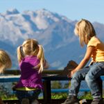 Drei junge Mädchen mit blondem Haar spielen zusammen auf einer Bank im Freien mit Bergen und blauem Himmel im Hintergrund.