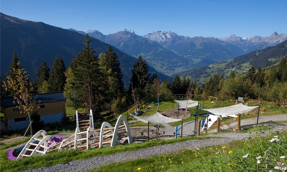 Spielplatz im Freien mit Klettergerüsten und schattenspendenden Vordächern in Hanglage, umgeben von Bäumen und mit Blick auf eine malerische Berglandschaft.