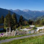 Spielplatz im Freien mit Klettergerüsten und schattenspendenden Vordächern in Hanglage, umgeben von Bäumen und mit Blick auf eine malerische Berglandschaft.
