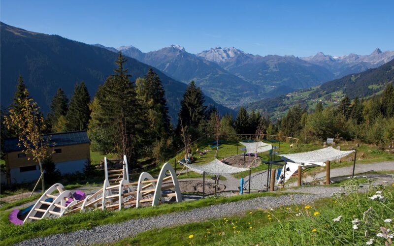 Spielplatz im Freien mit Klettergerüsten und schattenspendenden Vordächern in Hanglage, umgeben von Bäumen und mit Blick auf eine malerische Berglandschaft.