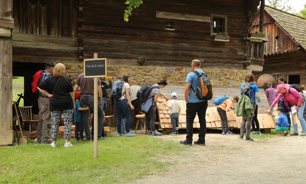 Eine Gruppe von Menschen, darunter Kinder und Erwachsene, versammelt sich vor einem rustikalen Holzgebäude und betrachtet die an der Gebäudewand aufgestellten Auslagen oder Aktivitäten.