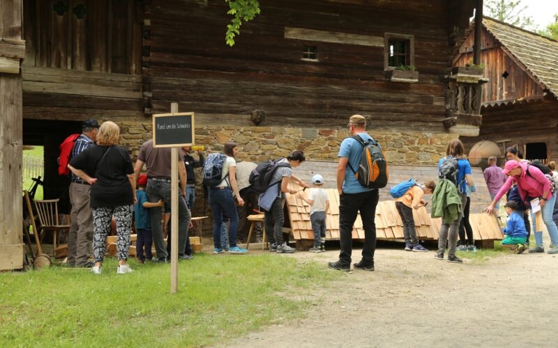 Eine Gruppe von Menschen, darunter Kinder und Erwachsene, versammelt sich vor einem rustikalen Holzgebäude und betrachtet die an der Gebäudewand aufgestellten Auslagen oder Aktivitäten.