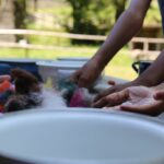 Mehrere Personen arbeiten mit bunten Wollbüscheln an einem Tisch im Freien, wobei im Vorder- und Hintergrund Schalen und Hände zu sehen sind.