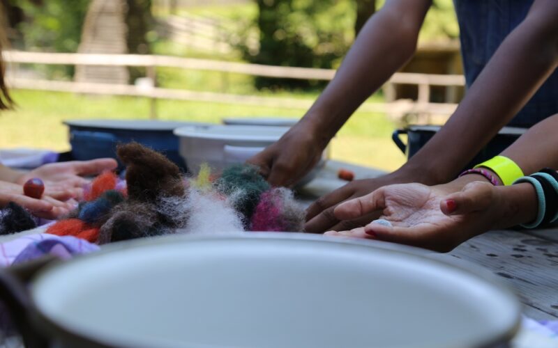 Mehrere Personen arbeiten mit bunten Wollbüscheln an einem Tisch im Freien, wobei im Vorder- und Hintergrund Schalen und Hände zu sehen sind.