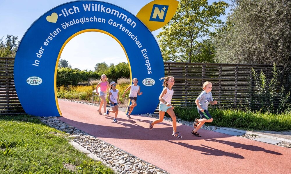Mehrere Kinder laufen auf einem roten Weg unter einem blauen Torbogen, auf dem "Herzlich Willkommen" steht und in deutscher Sprache für eine ökologische Gartenausstellung geworben wird. Im Hintergrund sind Bäume und Zäune zu sehen.
