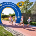 Mehrere Kinder laufen auf einem roten Weg unter einem blauen Torbogen, auf dem "Herzlich Willkommen" steht und in deutscher Sprache für eine ökologische Gartenausstellung geworben wird. Im Hintergrund sind Bäume und Zäune zu sehen.