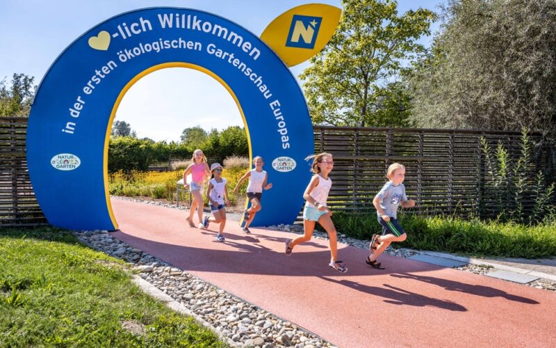 Mehrere Kinder laufen auf einem roten Weg unter einem blauen Torbogen, auf dem "Herzlich Willkommen" steht und in deutscher Sprache für eine ökologische Gartenausstellung geworben wird. Im Hintergrund sind Bäume und Zäune zu sehen.