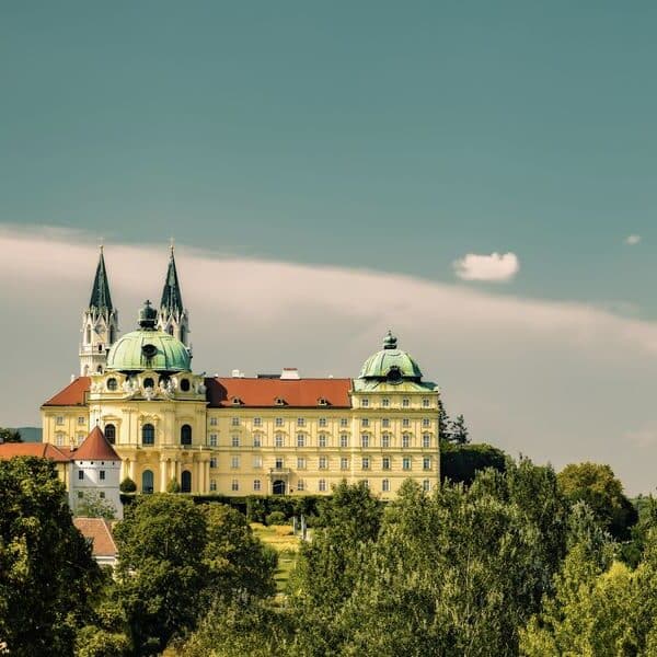 Ein großes historisches Gebäude mit grünen Kuppeln und Zwillingstürmen thront auf einem Hügel, umgeben von Bäumen unter einem teilweise bewölkten Himmel.
