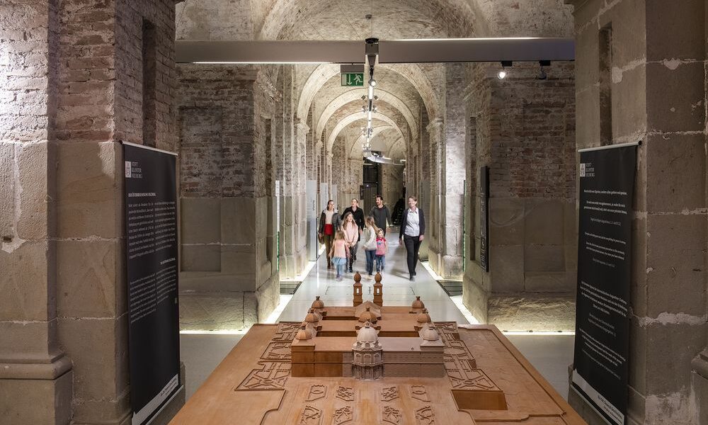 Eine Gruppe von Personen geht durch einen gemauerten Flur mit Steinsäulen; im Vordergrund ist ein großes hölzernes Architekturmodell zu sehen.