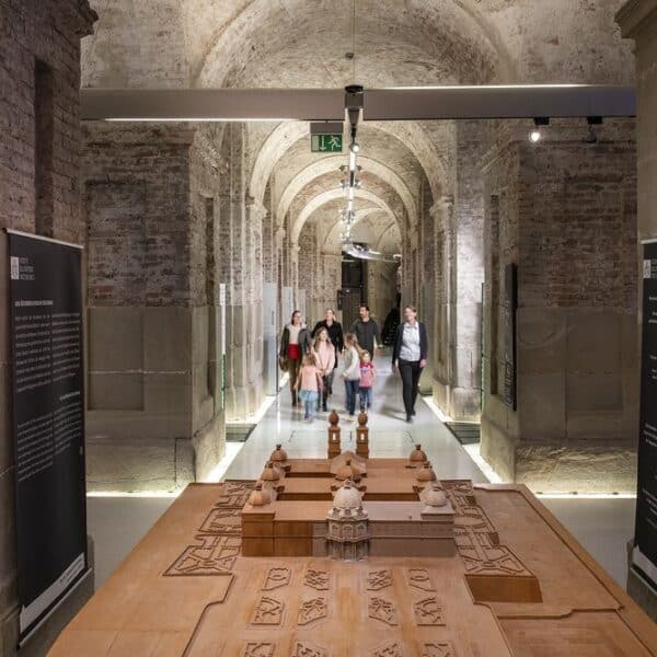 Eine Gruppe von Personen geht durch einen gemauerten Flur mit Steinsäulen; im Vordergrund ist ein großes hölzernes Architekturmodell zu sehen.