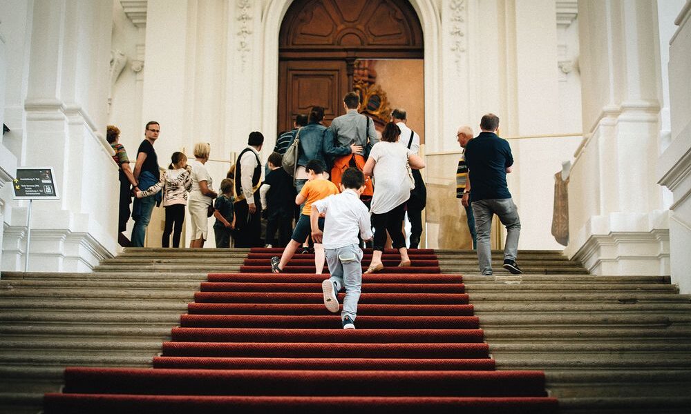 Eine Gruppe von Menschen, darunter auch Kinder, geht eine Treppe mit rotem Teppich hinauf zu einer großen Holztür in einem großen, weißen Gebäude.