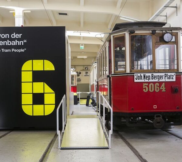 Eine alte rote Straßenbahn mit der Nummer 5064 steht in einem Innenraum neben einem großen schwarz-gelben Schild mit der Nummer 6 und einem zweisprachigen Zitat über Straßenbahnfahrer.