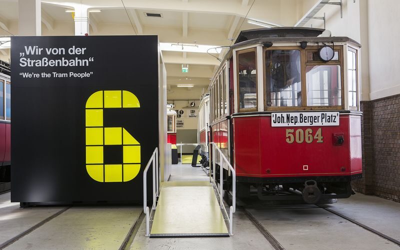 Eine alte rote Straßenbahn mit der Nummer 5064 steht in einem Innenraum neben einem großen schwarz-gelben Schild mit der Nummer 6 und einem zweisprachigen Zitat über Straßenbahnfahrer.