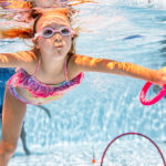 Ein kleines Kind mit Schwimmbrille schwimmt in einem Schwimmbecken unter Wasser und greift nach einem rosafarbenen Spielzeugring, während im Hintergrund andere Kinder zu sehen sind.