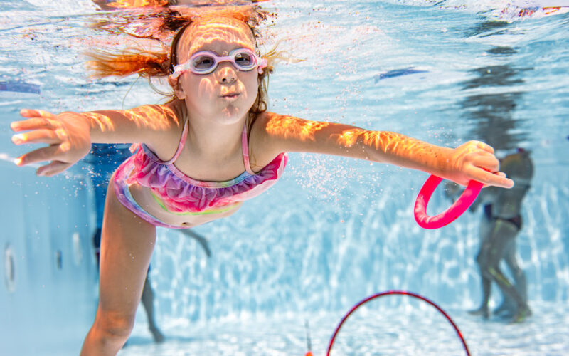 Ein kleines Kind mit Schwimmbrille schwimmt in einem Schwimmbecken unter Wasser und greift nach einem rosafarbenen Spielzeugring, während im Hintergrund andere Kinder zu sehen sind.