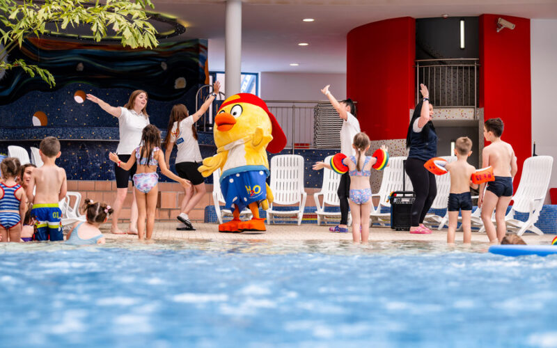 Kinder und Erwachsene in Badeanzügen stehen neben einem Hallenbad, einige tanzen mit einer Person in einem großen gelben Entenkostüm. Im Hintergrund sind weiße Stühle und bunte Wände zu sehen.