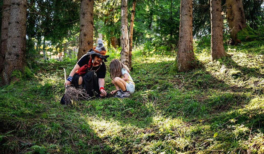 Ein Erwachsener und ein Kind hocken auf dem grasbewachsenen Waldboden und untersuchen bei Tageslicht zwischen hohen Bäumen etwas auf dem Boden.