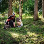 Ein Erwachsener und ein Kind hocken auf dem grasbewachsenen Waldboden und untersuchen bei Tageslicht zwischen hohen Bäumen etwas auf dem Boden.