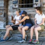 Drei Frauen sitzen auf einer Holzbank vor einem rustikalen Gebäude; eine von ihnen hält ein braunes Huhn im Arm, während die anderen zusehen und miteinander plaudern.