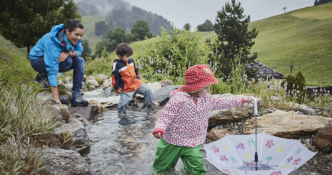 Ein Erwachsener und zwei Kinder spielen an einem seichten Bach in einer grasbewachsenen, bergigen Landschaft. Ein Kind hält einen aufgespannten Regenschirm ins Wasser, während die anderen zusehen.
