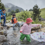 Ein Erwachsener und zwei Kinder spielen an einem seichten Bach in einer grasbewachsenen, bergigen Landschaft. Ein Kind hält einen aufgespannten Regenschirm ins Wasser, während die anderen zusehen.