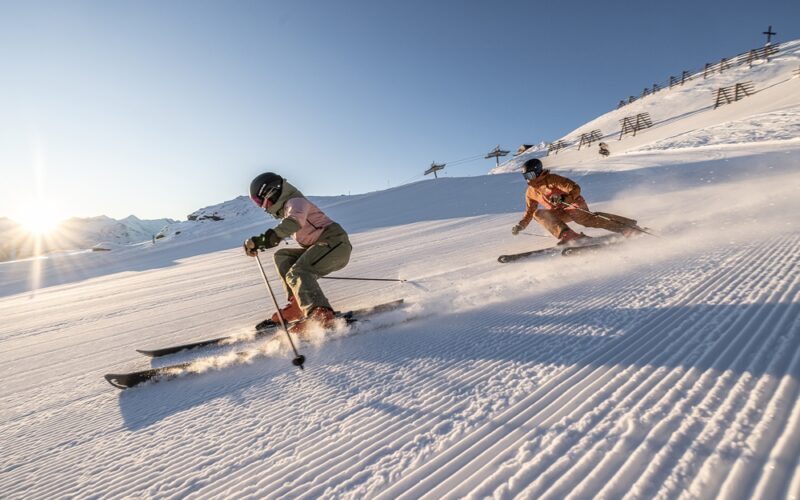 Zwei Personen in Winterausrüstung fahren auf einer präparierten, verschneiten Piste mit Sonnenlicht im Hintergrund und sichtbaren Bergen.