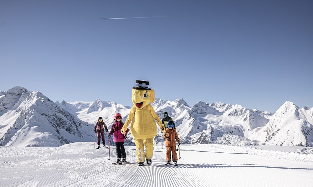 Eine Gruppe von Skifahrern, darunter eine Person in einem großen gelben Bärenkostüm, steht auf einer präparierten Piste mit schneebedeckten Bergen im Hintergrund.