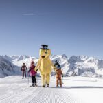 Eine Gruppe von Skifahrern, darunter eine Person in einem großen gelben Bärenkostüm, steht auf einer präparierten Piste mit schneebedeckten Bergen im Hintergrund.
