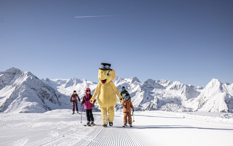 Eine Gruppe von Skifahrern, darunter eine Person in einem großen gelben Bärenkostüm, steht auf einer präparierten Piste mit schneebedeckten Bergen im Hintergrund.