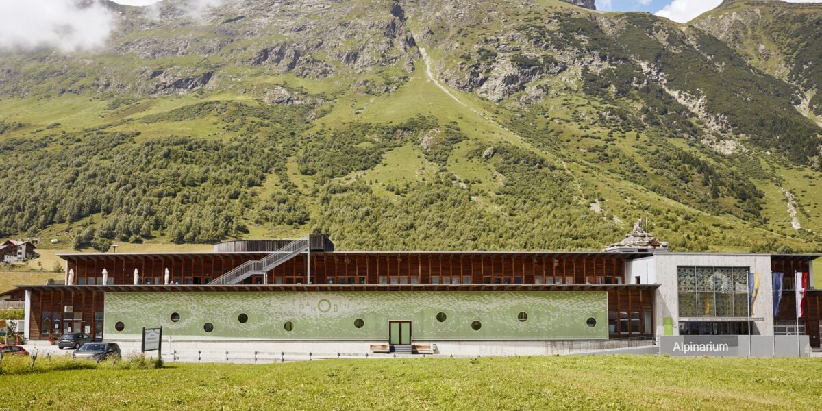 Ein modernes Gebäude mit der Aufschrift "Alpinarium" steht vor einem grünen Berghang unter einem teilweise bewölkten Himmel.