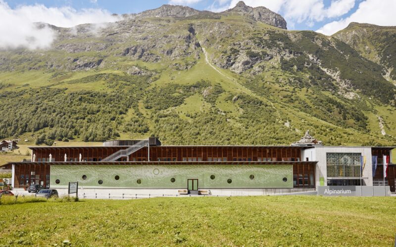 Ein modernes Gebäude mit der Aufschrift "Alpinarium" steht vor einem grünen Berghang unter einem teilweise bewölkten Himmel.