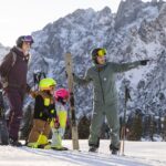 Zwei Erwachsene und zwei Kinder in Skiausrüstung stehen auf einem verschneiten Berghang mit Skiern, während ein Erwachsener in die Ferne zeigt. Im Hintergrund sind schneebedeckte Berge zu sehen.