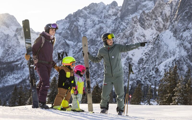 Zwei Erwachsene und zwei Kinder in Skiausrüstung stehen auf einem verschneiten Berghang mit Skiern, während ein Erwachsener in die Ferne zeigt. Im Hintergrund sind schneebedeckte Berge zu sehen.