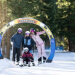 Vier Personen in Skiausrüstung posieren unter einem bunten "Funslope"-Bogen auf einer verschneiten, von Bäumen umgebenen Piste.