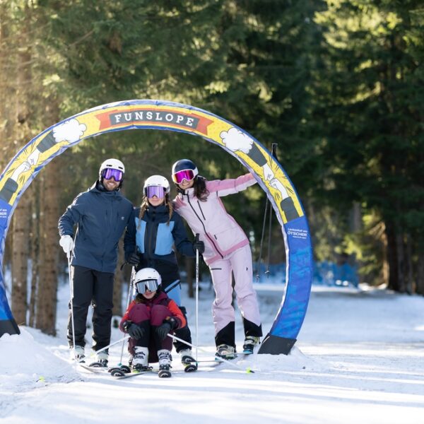 Vier Personen in Skiausrüstung posieren unter einem bunten "Funslope"-Bogen auf einer verschneiten, von Bäumen umgebenen Piste.