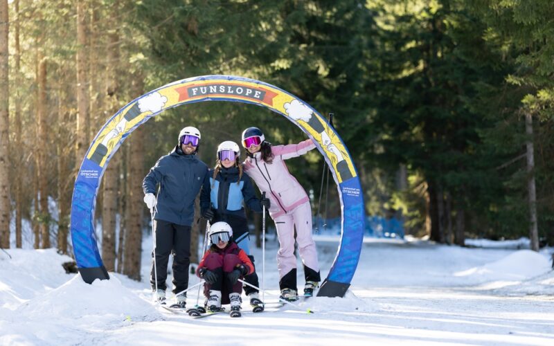 Vier Personen in Skiausrüstung posieren unter einem bunten "Funslope"-Bogen auf einer verschneiten, von Bäumen umgebenen Piste.