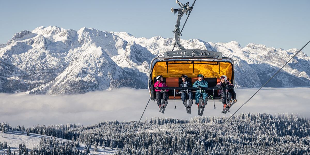 Vier Skifahrer fahren mit einem gelben Skilift über schneebedeckte Bäume, mit Bergen und einem bewölkten Himmel im Hintergrund.