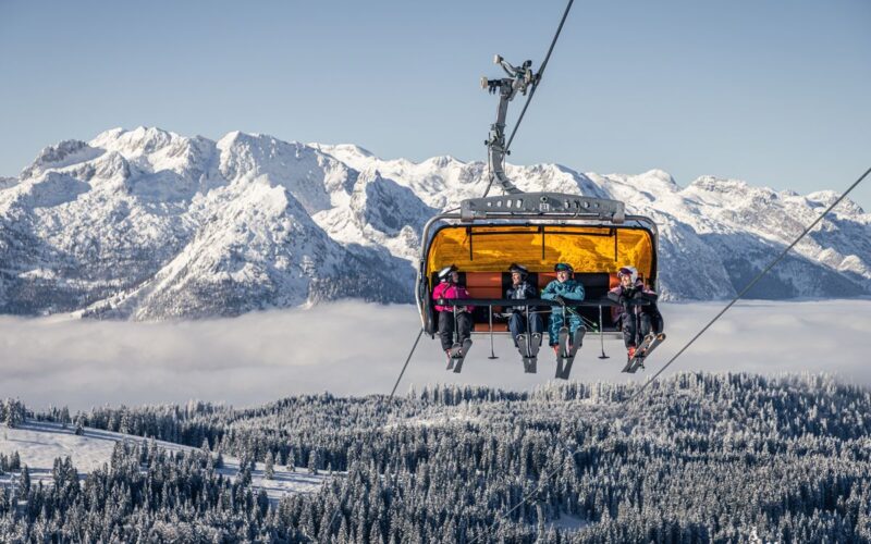 Vier Skifahrer fahren mit einem gelben Skilift über schneebedeckte Bäume, mit Bergen und einem bewölkten Himmel im Hintergrund.