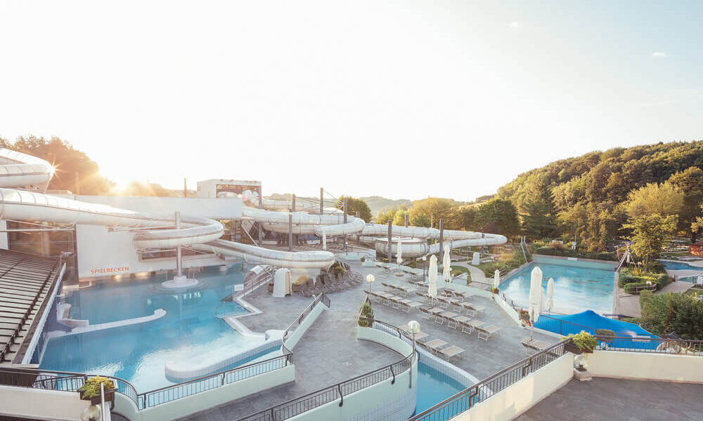 Wasserpark im Freien mit mehreren Becken, Wasserrutschen, Liegestühlen und Sonnenschirmen, an einem sonnigen Tag umgeben von Bäumen.