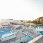 Wasserpark im Freien mit mehreren Becken, Wasserrutschen, Liegestühlen und Sonnenschirmen, an einem sonnigen Tag umgeben von Bäumen.