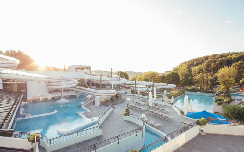 Wasserpark im Freien mit mehreren Becken, Wasserrutschen, Liegestühlen und Sonnenschirmen, an einem sonnigen Tag umgeben von Bäumen.