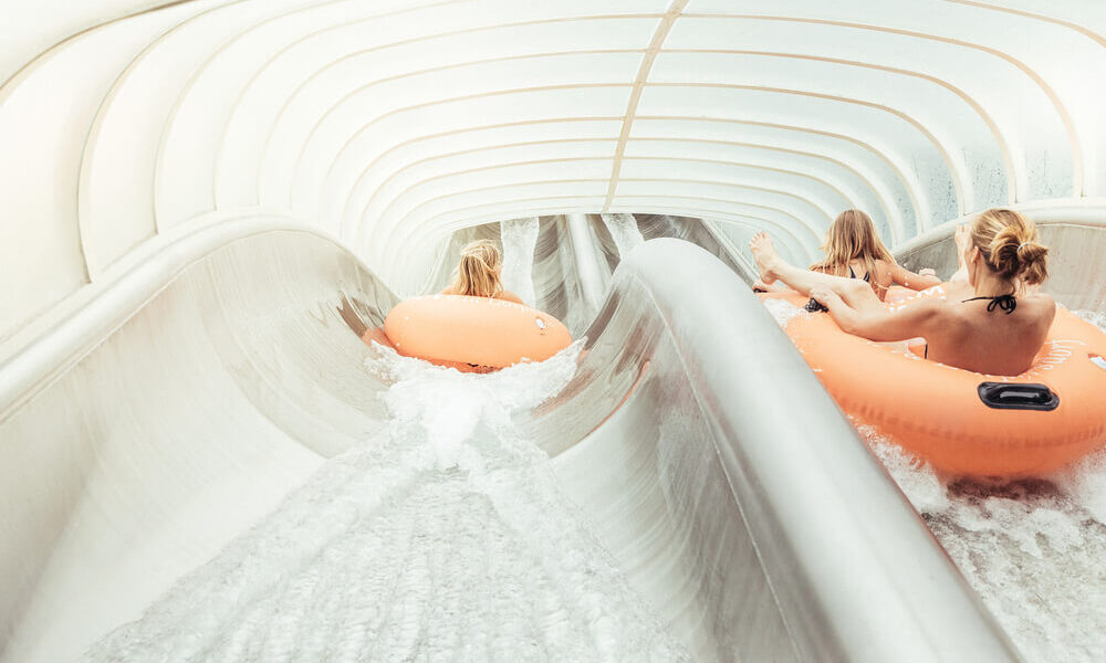 Drei Personen fahren mit orangefarbenen Schläuchen eine überdachte Wasserrutsche hinunter, wobei das Wasser im Tunnel um sie herumspritzt.