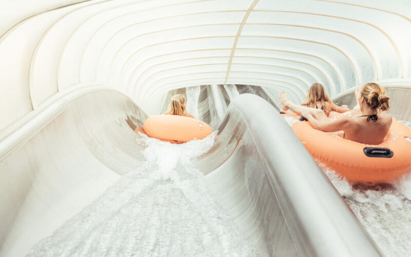 Drei Personen fahren mit orangefarbenen Schläuchen eine überdachte Wasserrutsche hinunter, wobei das Wasser im Tunnel um sie herumspritzt.
