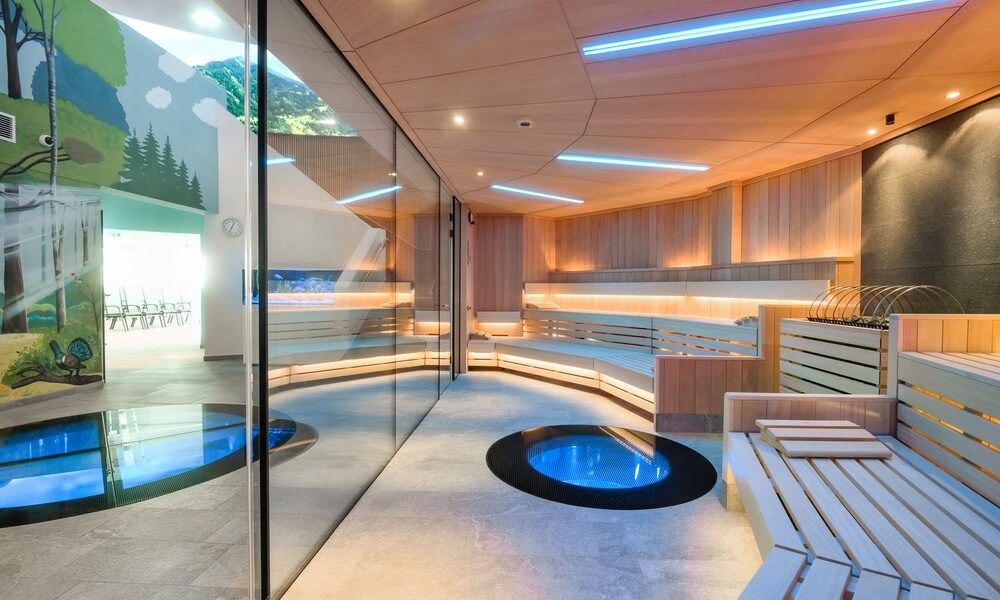 Moderne Sauna mit Holzbänken, blauer LED-Beleuchtung, zwei glasgeschützten runden Becken im Boden und einer Glaswand, die sie von einem mit Wandmalereien bemalten Raum im Freien trennt.