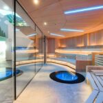 Moderne Sauna mit Holzbänken, blauer LED-Beleuchtung, zwei glasgeschützten runden Becken im Boden und einer Glaswand, die sie von einem mit Wandmalereien bemalten Raum im Freien trennt.