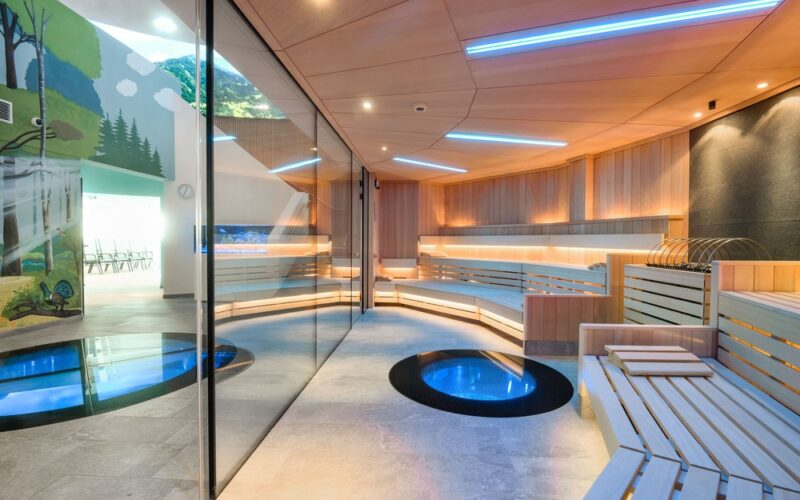 Moderne Sauna mit Holzbänken, blauer LED-Beleuchtung, zwei glasgeschützten runden Becken im Boden und einer Glaswand, die sie von einem mit Wandmalereien bemalten Raum im Freien trennt.