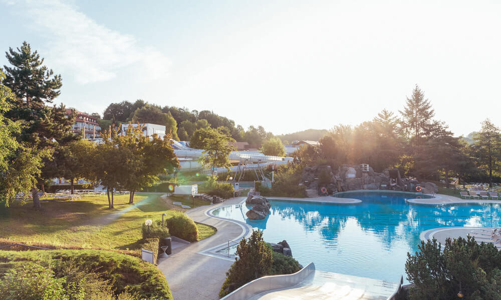 Das Freibad ist von Bäumen und Grün umgeben, der Himmel ist klar und die Sonne scheint auf die Landschaft.