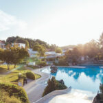 Das Freibad ist von Bäumen und Grün umgeben, der Himmel ist klar und die Sonne scheint auf die Landschaft.