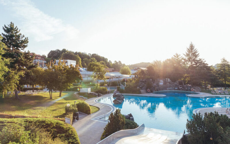 Das Freibad ist von Bäumen und Grün umgeben, der Himmel ist klar und die Sonne scheint auf die Landschaft.