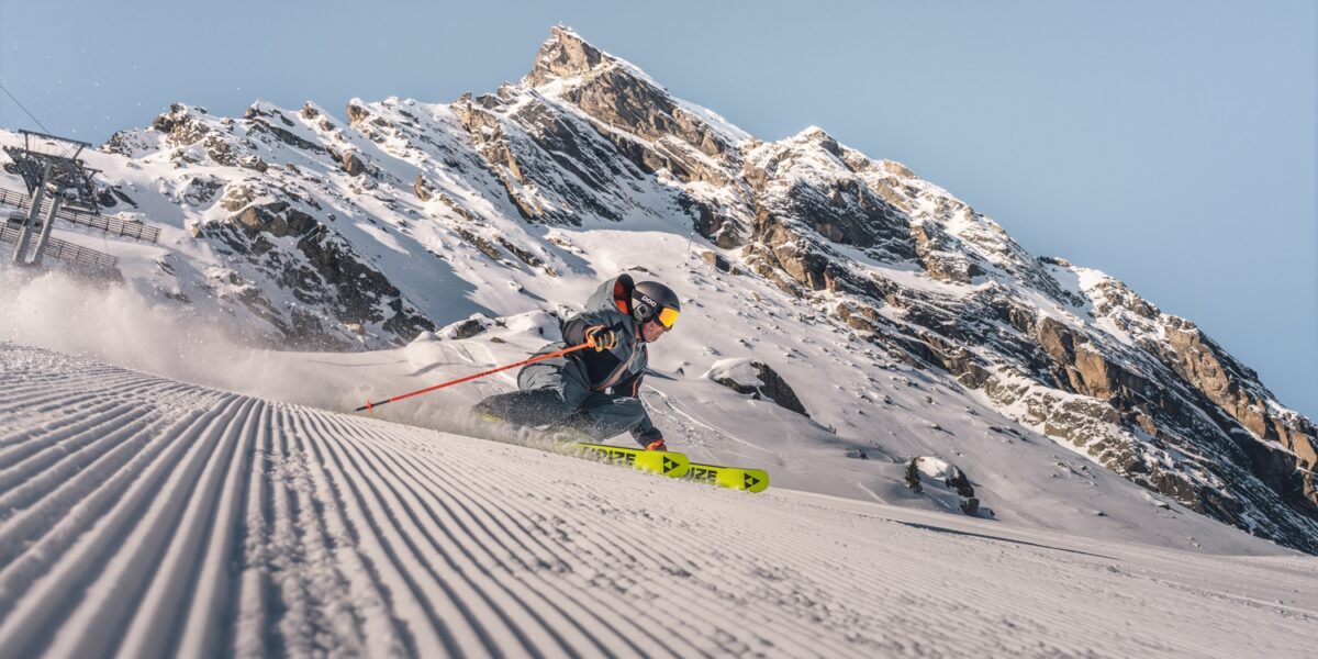 Ein Skifahrer in Winterausrüstung fährt eine frisch präparierte Piste hinunter, während im Hintergrund ein schneebedeckter Berg zu sehen ist.